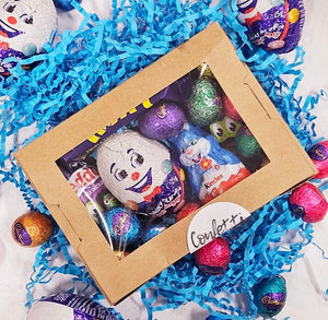 A festive Easter-themed gift box filled with assorted chocolates. The box has a clear window on top, displaying a variety of colorful foil-wrapped Easter eggs, a Kinder bunny, Cadbury chocolates, and a smiling Humpty Dumpty chocolate. The box is surrounded by bright blue shredded paper and more foil-wrapped eggs scattered around