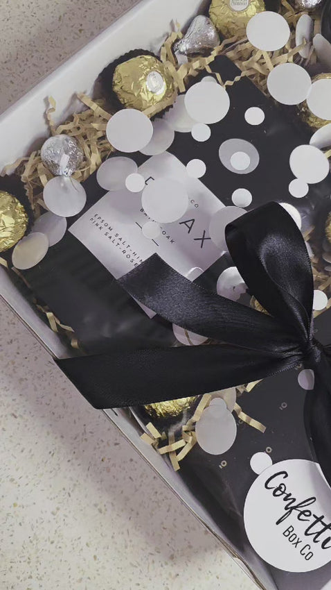 A Perth-made Soak & Sip self care gift box with bath soak, chocolates and wine, packed in a white box with black ribbon and confetti.