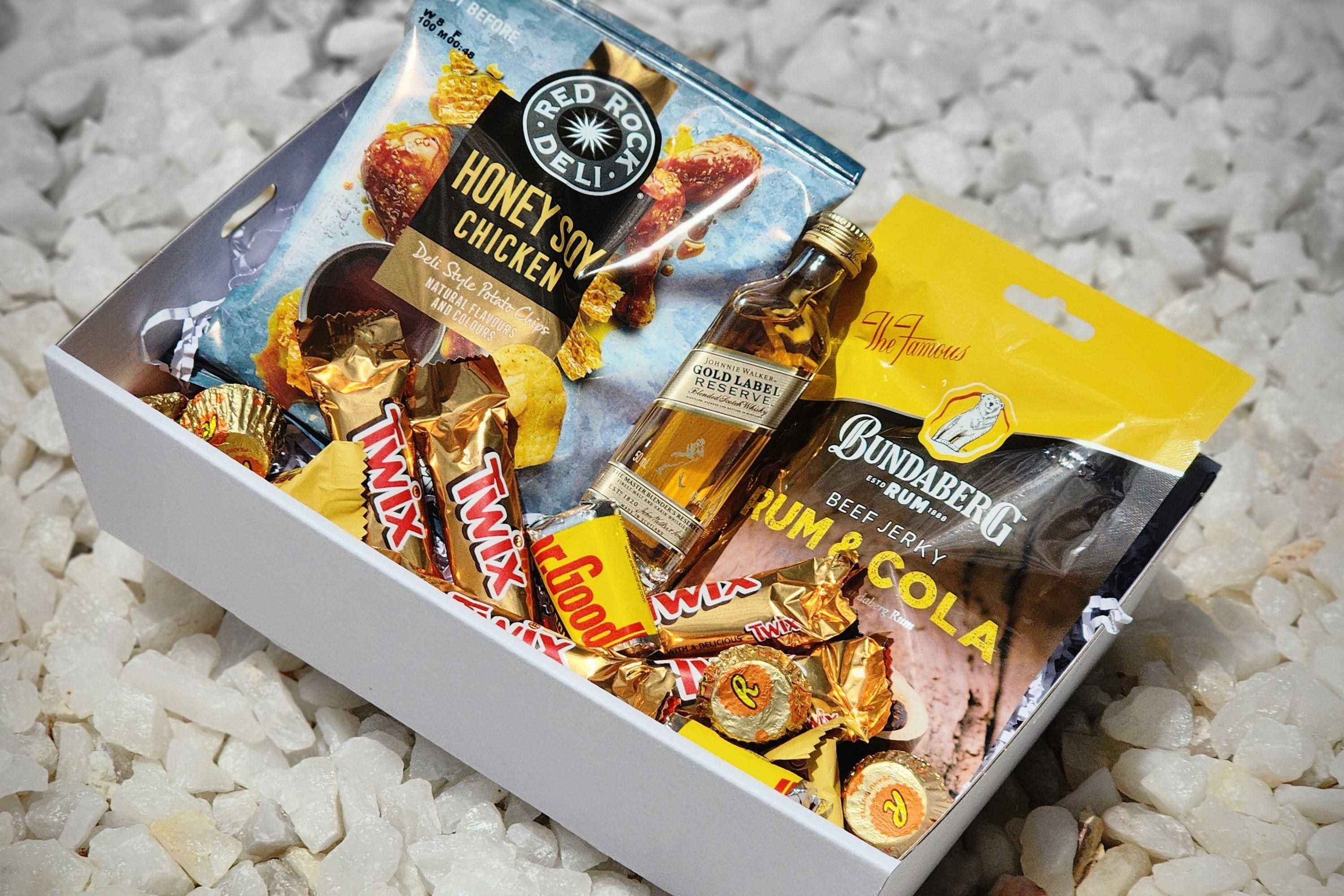 Gold Label Johnnie Walker Mini & Treats: The perfect small gift for scotch lovers. Includes 50ml Gold Label Johnnie Walker, Red Rock Deli Honey Soy Chips, Dr Proctor’s Beef Jerky, Mini Twix, Hershey’s, Reese’s Peanut Butter Cups, and Toblerone chocolates, all elegantly packed in a gift box